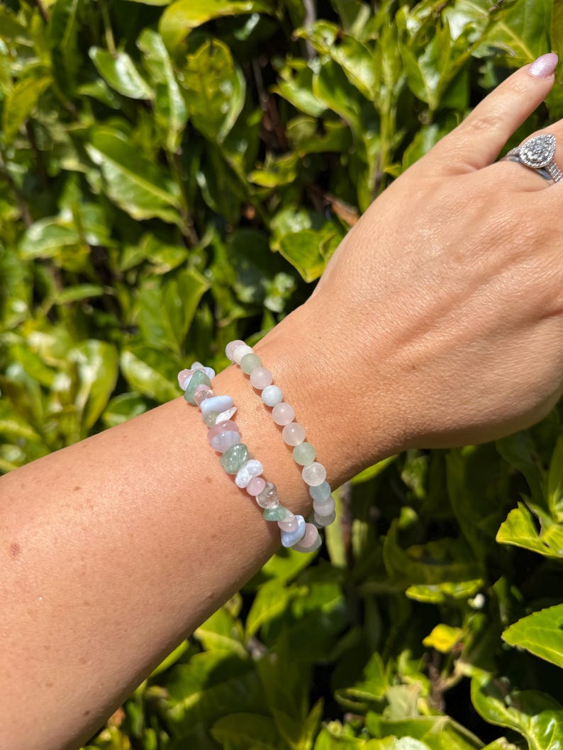 May include: Two gemstone bracelets are displayed on a wrist. One bracelet features small, round beads in shades of pink and green. The other bracelet has a mix of irregular, pastel-colored stones. The background is a lush green bush.