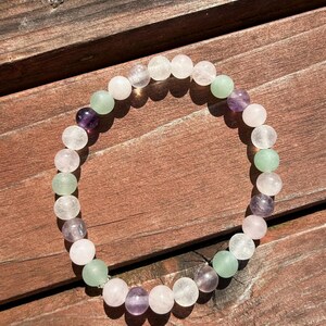 May include: A beaded bracelet featuring a variety of colorful gemstones. The bracelet includes clear, light green, purple, and pink beads. The bracelet is displayed on a wooden surface, showcasing the natural colors of the stones.