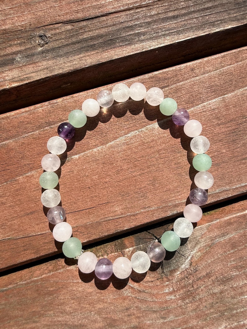 May include: A beaded bracelet featuring a variety of colorful gemstones. The bracelet includes clear, light green, purple, and pink beads. The bracelet is displayed on a wooden surface, showcasing the natural colors of the stones.