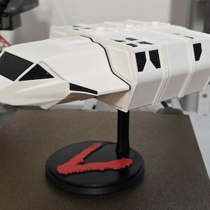 Peut inclure: Maquette blanche de vaisseau spatial avec des détails noirs et des marquages rouges, montée sur un socle noir avec un motif en forme de V rouge. Le vaisseau spatial a un design angulaire et futuriste.