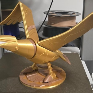Peut inclure: Une sculpture d'oiseau doré aux ailes déployées, posée sur une base circulaire. L'oiseau présente un design détaillé, avec un bec et des accents bruns. La sculpture est probablement imprimée en 3D et repose sur une surface sombre.