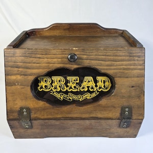 May include: A brown wooden bread box with a hinged front door featuring a central glass window. The glass displays the yellow text BREAD and the phrase MADE WITH TENDER LOVING CARE. The box includes a small dark knob and metal hinges.