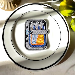 May include: An embroidered patch depicting a can of sardines. The patch features a gray border, with a blue and orange interior. Three fish are visible inside the can. The patch is placed on a white plate with a black rim.