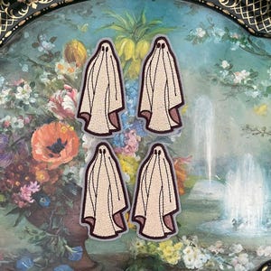 May include: Four beige ghost patches with brown trim are arranged on a decorative tray with a floral painting. The patches have a simple ghost design with a rounded bottom and eye holes. The tray has a black and gold ornate border.