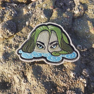 May include: Embroidered patch depicting a face with green hair, blue eyes, and a blue water-like border. The patch is set against a textured, light brown background, reminiscent of a coastal environment.
