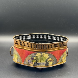 May include: An oval-shaped, vintage metal planter with a decorative floral design. The planter features a red and gold color scheme, with a detailed rim and handles. The floral design includes potted plants.