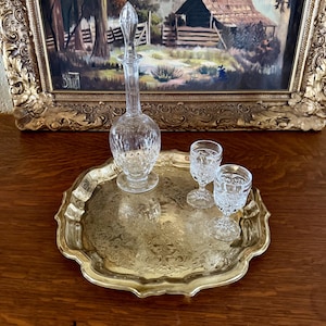 May include: A round brass tray with an ornate embossed floral pattern and scalloped edges. Resting on the tray is a clear glass decanter with a matching stopper and two small clear glass stemmed goblets, all featuring a cut glass pattern.