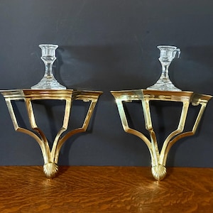 May include: A pair of gold-toned metal wall sconce shelves featuring a geometric frame with a shell-shaped base detail. Each shelf supports a clear, faceted glass candle holder with a wide rim and a tapered stem, designed for holding taper candles.