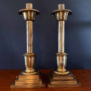May include: Two brass candlesticks with a classic design. Each candlestick has a square base, a cylindrical stem, and a flared top. The brass has a polished finish, reflecting light. The candlesticks are displayed on a wooden surface against a dark blue background.