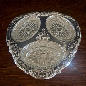 May include: A silver-toned metal serving tray featuring three oval-shaped clear glass inserts with a sunburst pattern. The metal frame has an ornate, embossed border with floral and scroll details, including a central floral accent piece.