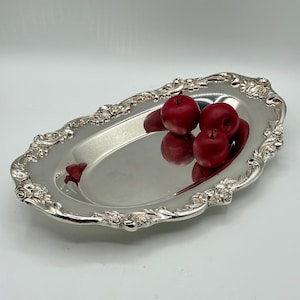 May include: An oval silver-toned serving tray with a polished reflective surface and an ornate raised border featuring detailed floral and scrollwork patterns. Three small, round, deep red decorative fruits rest inside the tray.