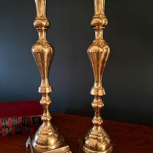 May include: Two ornate, gold-colored candlesticks with a square base and a tall, slender body. The candlesticks have a flared top to hold the candles. The background is a dark blue wall and a wooden table.
