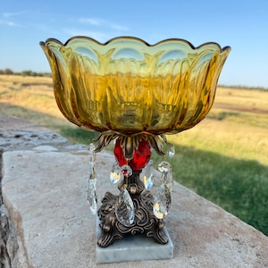 May include: A decorative amber glass bowl with a scalloped rim, supported by a bronze-toned metal base featuring intricate scrollwork, a central red glass bead, and dangling clear crystal teardrop prisms, all set on a square white marble base.