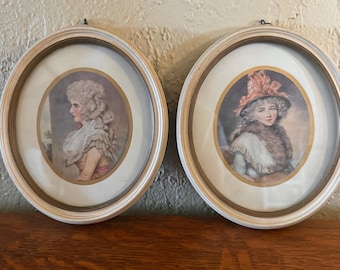 May include: Two oval wall frames with light-colored wooden borders, each containing a portrait print. The left print shows a person in profile with a white ruffled collar, while the right print depicts a person wearing a hat and a fur-trimmed garment.