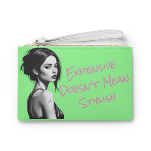 May include: A green clutch purse with a white zipper and a white strap. The purse features a black and white illustration of a woman with the text "Expensive doesn't mean stylish" written in pink.