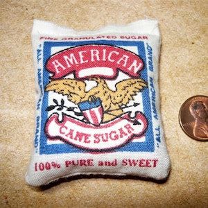 May include: A small, white fabric bag with red, white, and blue embroidery. The bag is shaped like a pillow and has the words "American Cane Sugar" and "100% Pure and Sweet" printed on it.