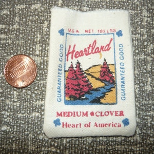 May include: A white fabric bag with the text "Heartland" in red and blue lettering. The bag also has the text "U.S.A. Net 100 Lbs. Guaranteed Good" and "Medium Clover Heart of America" printed on it. The bag features a colorful illustration of two trees and a river.