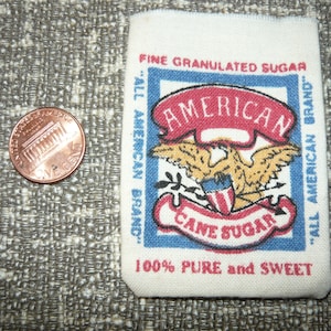 May include: A white fabric label with blue and red trim. The label features an eagle with a shield and the text "American Cane Sugar" in a blue banner. The text "Fine Granulated Sugar" and "All American Brand" are printed on the label.