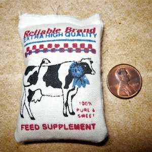Puede incluir: Una bolsa de tela blanca con ribetes rojos y azules. La bolsa tiene el texto "Reliable Brand Extra High Quality" y una ilustración en blanco y negro de una vaca. La bolsa también tiene el texto "Feed Supplement" y "100% Pure & Sweet".