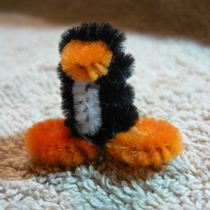 以下が含まれることがあります： パイプクリーナーで作った黒とオレンジのペンギン。ペンギンは白いふわふわの表面に立っています。