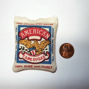 May include: A small white fabric bag with a red, white, and blue design. The bag has the words "American Cane Sugar" printed on it. The bag is about the size of a penny.