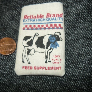 May include: A white bag with red and blue checkered trim and a black and white cow illustration. The bag has the text "Reliable Brand Extra High Quality" and "Feed Supplement" printed on it. The bag also has the text "100% Pure & Sweet" printed on it.