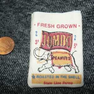 May include: A small, rectangular fabric bag with the words "FRESH GROWN" and "JUMBO PEANUTS" printed on it. An illustration of an elephant is featured. A penny is next to the bag.