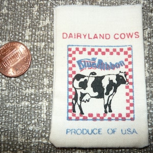 May include: White fabric patch with red and blue checkered border. The patch features a black and white cow with the text "Dairyland Cows" and "Blue Ribbon" in a blue banner. The text "Produce of USA" is printed below the cow.