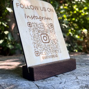 May include: Wooden Instagram sign with a QR code and the text "FOLLOW US ON Instagram". The sign is light-colored with a dark brown base. The Instagram logo is visible within the QR code. The sign is displayed outdoors.