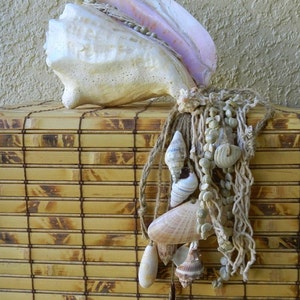 May include: A large white seashell with a pink interior, hanging from a woven rope with smaller seashells and beads. The seashells are hanging from a bamboo mat.