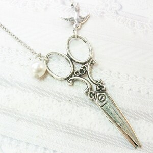 May include: A silver-colored necklace featuring a detailed scissor pendant with a pearl and a small bird charm. The scissors have ornate handles and sharp blades. The necklace hangs on a delicate chain, perfect for a unique accessory.