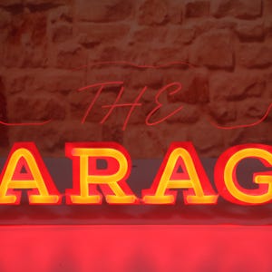 K&ouml;nnte beinhalten: Ein Neon-Schild mit der Aufschrift "THE GARAGE" in Rot und Gelb. Das Wort "GARAGE" ist in gro&szlig;en Blockbuchstaben mit gelbem Leuchten geschrieben, w&auml;hrend "THE" in einer d&uuml;nneren, roten Schrift gehalten ist. Der Hintergrund ist eine strukturierte Backsteinmauer.