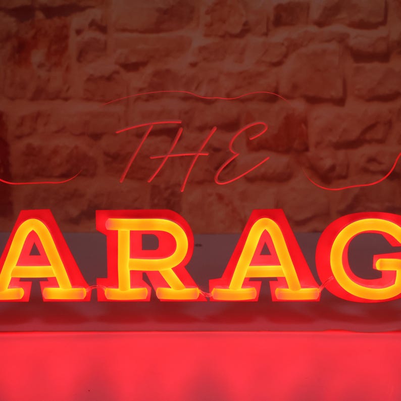 K&ouml;nnte beinhalten: Ein Neon-Schild mit der Aufschrift "THE GARAGE" in Rot und Gelb. Das Wort "GARAGE" ist in gro&szlig;en Blockbuchstaben mit gelbem Leuchten geschrieben, w&auml;hrend "THE" in einer d&uuml;nneren, roten Schrift gehalten ist. Der Hintergrund ist eine strukturierte Backsteinmauer.
