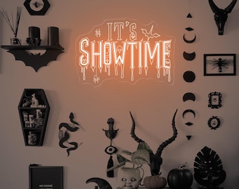 Op de afbeelding: Een Halloween-thema wanddecoratie met een neonbord met de tekst "IT'S SHOWTIME". De display bevat een vleermuisvormige plank, een doodskistvormige plank, een zwarte schedel en diverse andere spookachtige decoraties. Het kleurenschema is voornamelijk zwart en oranje.