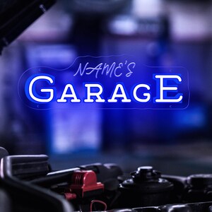 K&ouml;nnte beinhalten: Blaues Neon-Schild mit dem Text "NAME'S GARAGE" in wei&szlig;er Schrift. Das Schild leuchtet vor einem verschwommenen Hintergrund, mit dem Motor eines Autos im Vordergrund.