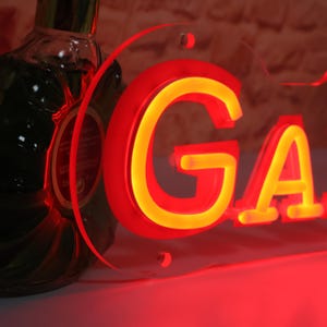 K&ouml;nnte beinhalten: Ein Neonschild mit den Buchstaben "GA" in Rot mit gelbem Schein. Das Schild ist auf einer transparenten Acrylplatte montiert. Eine dunkle Flasche ist teilweise im Hintergrund zu sehen.
