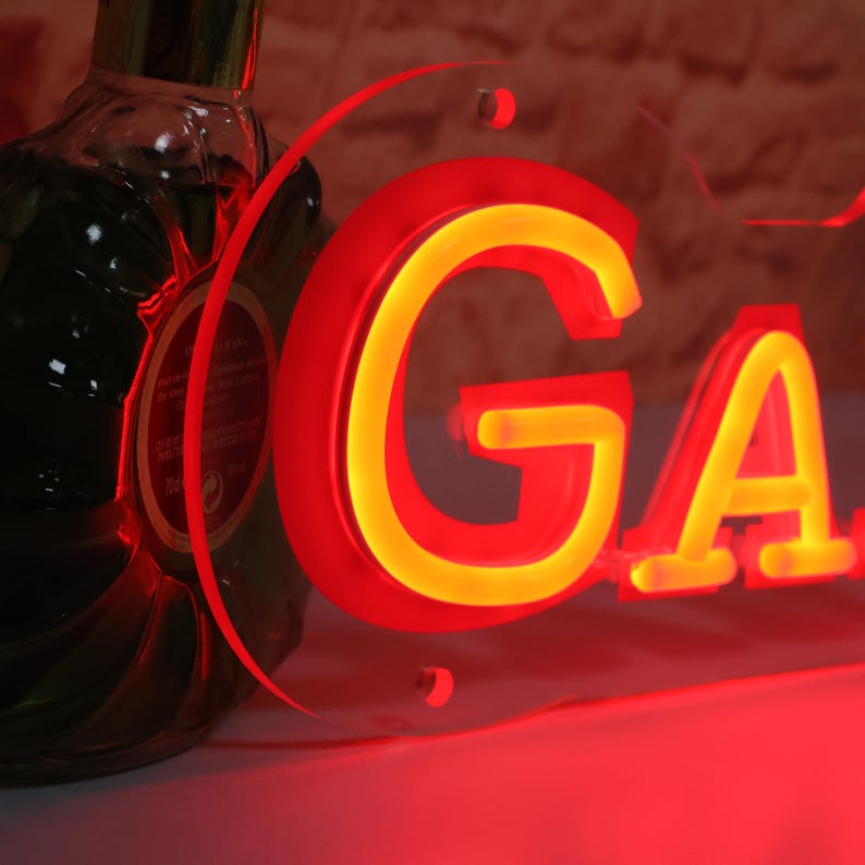 K&ouml;nnte beinhalten: Ein Neonschild mit den Buchstaben "GA" in Rot mit gelbem Schein. Das Schild ist auf einer transparenten Acrylplatte montiert. Eine dunkle Flasche ist teilweise im Hintergrund zu sehen.