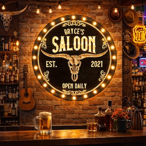 Könnte beinhalten: Ein rundes, beleuchtetes Schild für "Bryce's Saloon" mit einem Stierschädel-Motiv. Das Schild ist von warmweißen Lichtern umgeben und an einer Backsteinmauer befestigt. Die Bar ist mit Getränken und einem Bierkrug bestückt.