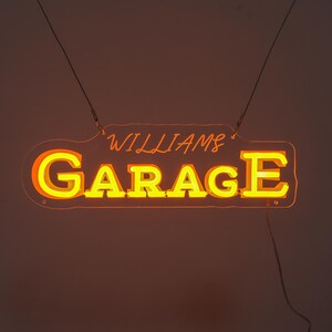 K&ouml;nnte beinhalten: Ein Neon-Schild mit der Aufschrift "WILLIAMS GARAGE" in Gelb und Orange. Das rechteckige Schild mit abgerundeten Ecken wird von d&uuml;nnen Dr&auml;hten gehalten. Der Hintergrund ist ein warmes, dunkles Orange.