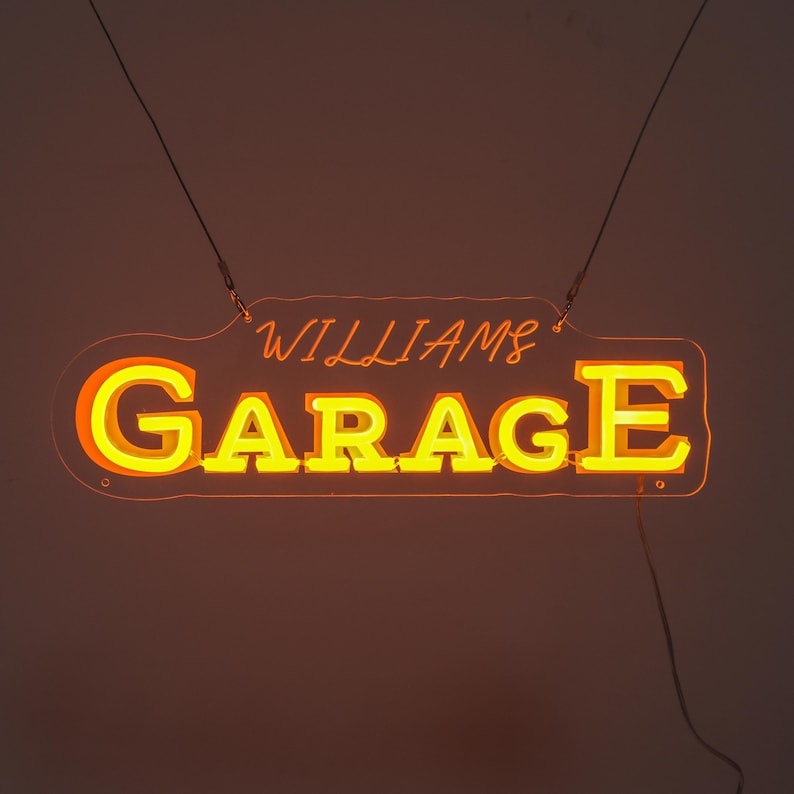 K&ouml;nnte beinhalten: Ein Neon-Schild mit der Aufschrift "WILLIAMS GARAGE" in Gelb und Orange. Das rechteckige Schild mit abgerundeten Ecken wird von d&uuml;nnen Dr&auml;hten gehalten. Der Hintergrund ist ein warmes, dunkles Orange.