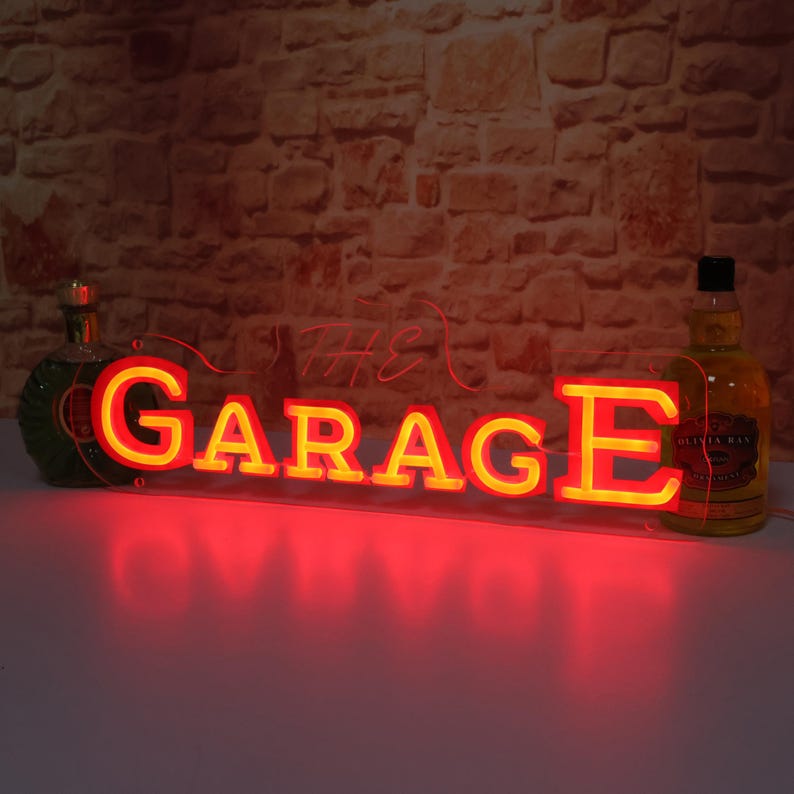 K&ouml;nnte beinhalten: Ein Neonschild mit dem Wort "GARAGE" in gelben Buchstaben mit roter Umrandung. Das Wort "THE" steht in kursiver Schrift dar&uuml;ber. Das Schild ist beleuchtet und steht vor einer Backsteinmauer. Flaschen stehen daneben.