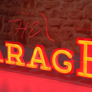K&ouml;nnte beinhalten: Rotes Neon-Schild mit dem Wort "GARAGE" in gro&szlig;en, leuchtenden Buchstaben. Das Wort "THE" steht dar&uuml;ber in Schreibschrift. Das Schild hat einen gelben Schein in den Buchstaben, vor einer Backsteinmauer.