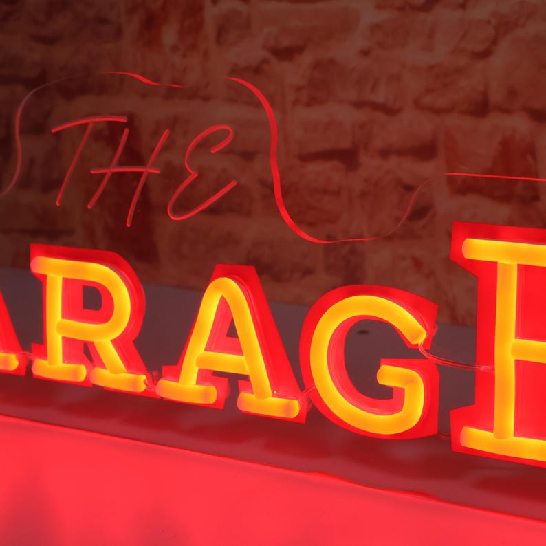 K&ouml;nnte beinhalten: Rotes Neon-Schild mit dem Wort "GARAGE" in gro&szlig;en, leuchtenden Buchstaben. Das Wort "THE" steht dar&uuml;ber in Schreibschrift. Das Schild hat einen gelben Schein in den Buchstaben, vor einer Backsteinmauer.