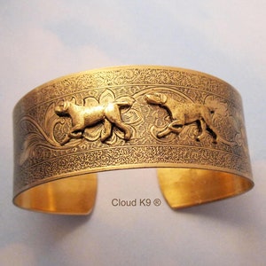 May include: A wide, gold-coloured cuff bracelet with an embossed floral pattern and two running dog figures. The bracelet has a polished finish and the text "Cloud K9 ®" is visible on the bottom.