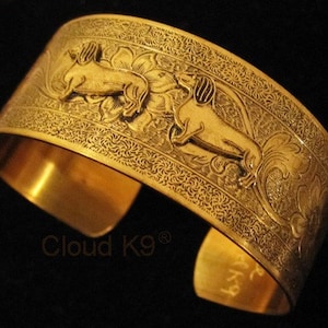 May include: Gold-toned metal cuff bracelet with an engraved design of two dachshunds. The bracelet has a detailed floral pattern and the words "Cloud K9" are engraved on the bottom.