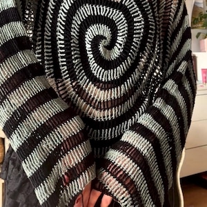 May include: A long-sleeved crochet jumper featuring a prominent black and light green spiral pattern on the front. The garment is constructed with an open-knit texture, showcasing alternating horizontal stripes of black and light green on the sleeves.