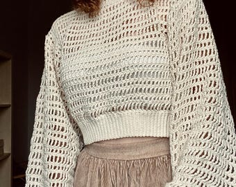 May include: A cream-colored crochet sweater with a loose fit and long sleeves. The sweater has a delicate open-stitch pattern and a cropped length.