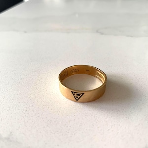 Peut inclure: Une bague dorée avec un symbole triangulaire noir et du texte gravé. La bague est posée sur une surface blanche. La bague est de style alliance, avec une surface plate.