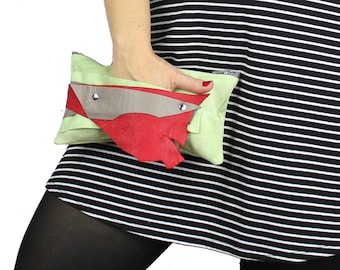 May include: A green, red, and gray leather clutch purse with a silver zipper. The purse is held by a person wearing a black and white striped dress.