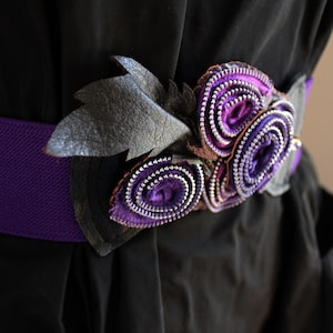 Può includere: Una cintura in tessuto viola con un fiore decorativo fatto con cerniere e una foglia di cuoio nero.